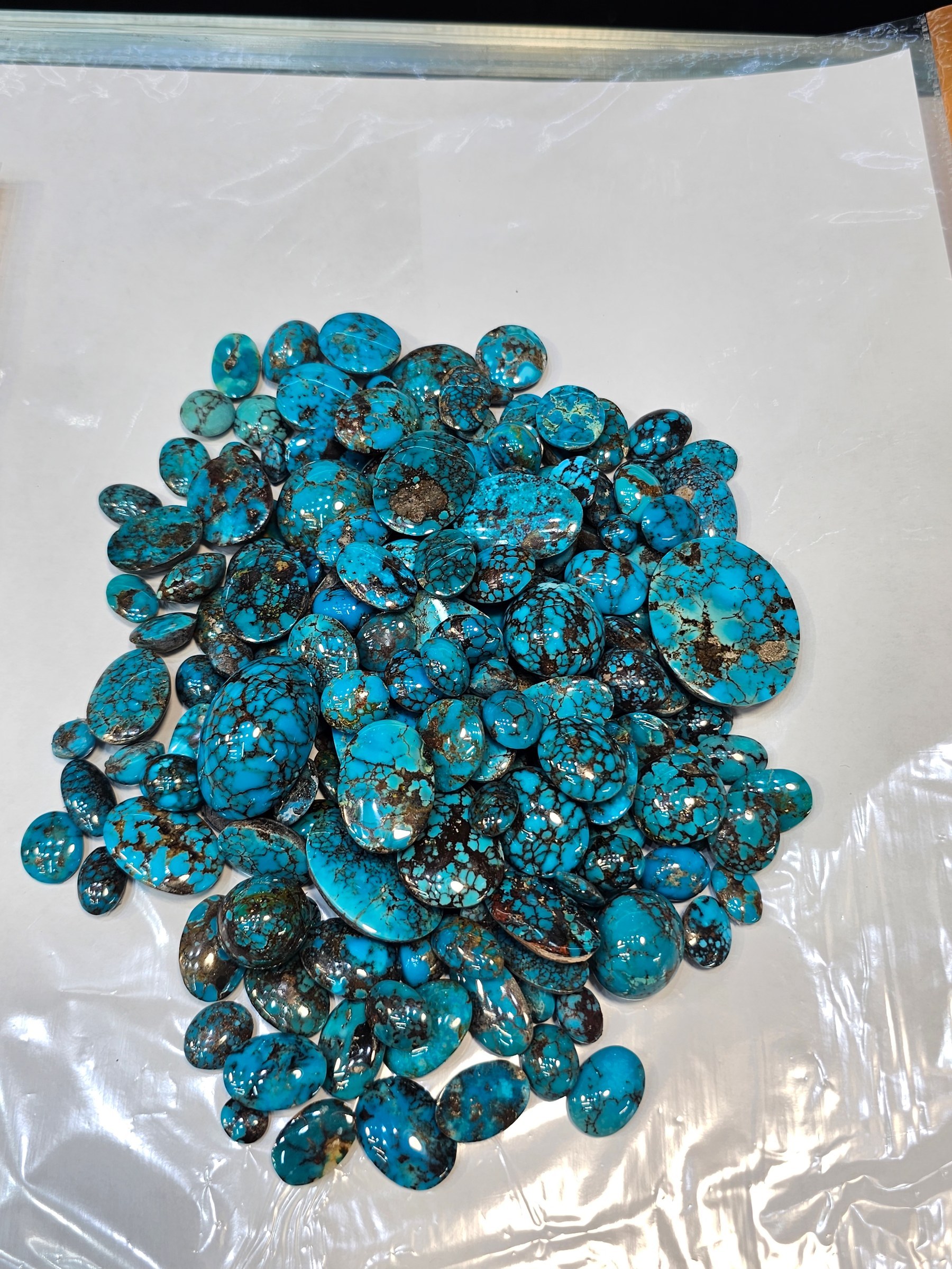A mound of loose polished turquoise cabochons in blue with dark matrix on a white sheet.
